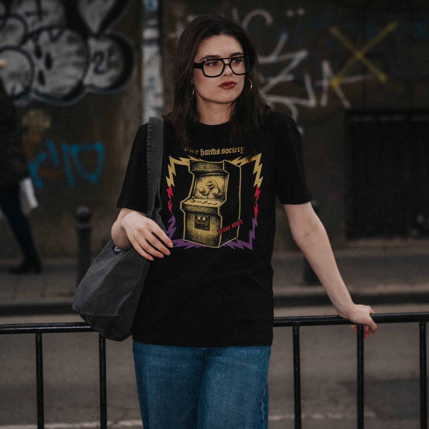Woman wearing a black t-shirt with a graphic design, standing against a graffiti-covered wall.